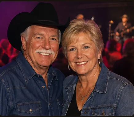 Smiling couple in denim shirts, one in a black cowboy hat, at a live country music show