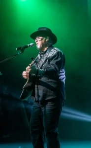 SST_CMC_Guitar_v2a Guitarist in a black cowboy hat and embroidered western shirt singing and playing under green stage lights